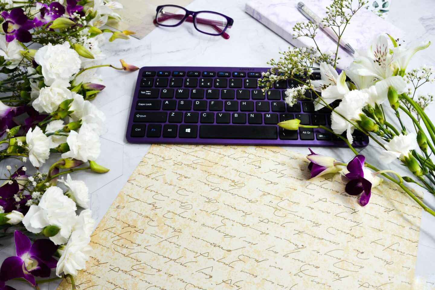 Landscape-Grey-marbled-background-with-calligraphy-parchment-paper-and-central-puple-keyboard-with-white purple-flowers-on-it-purple-glasses-